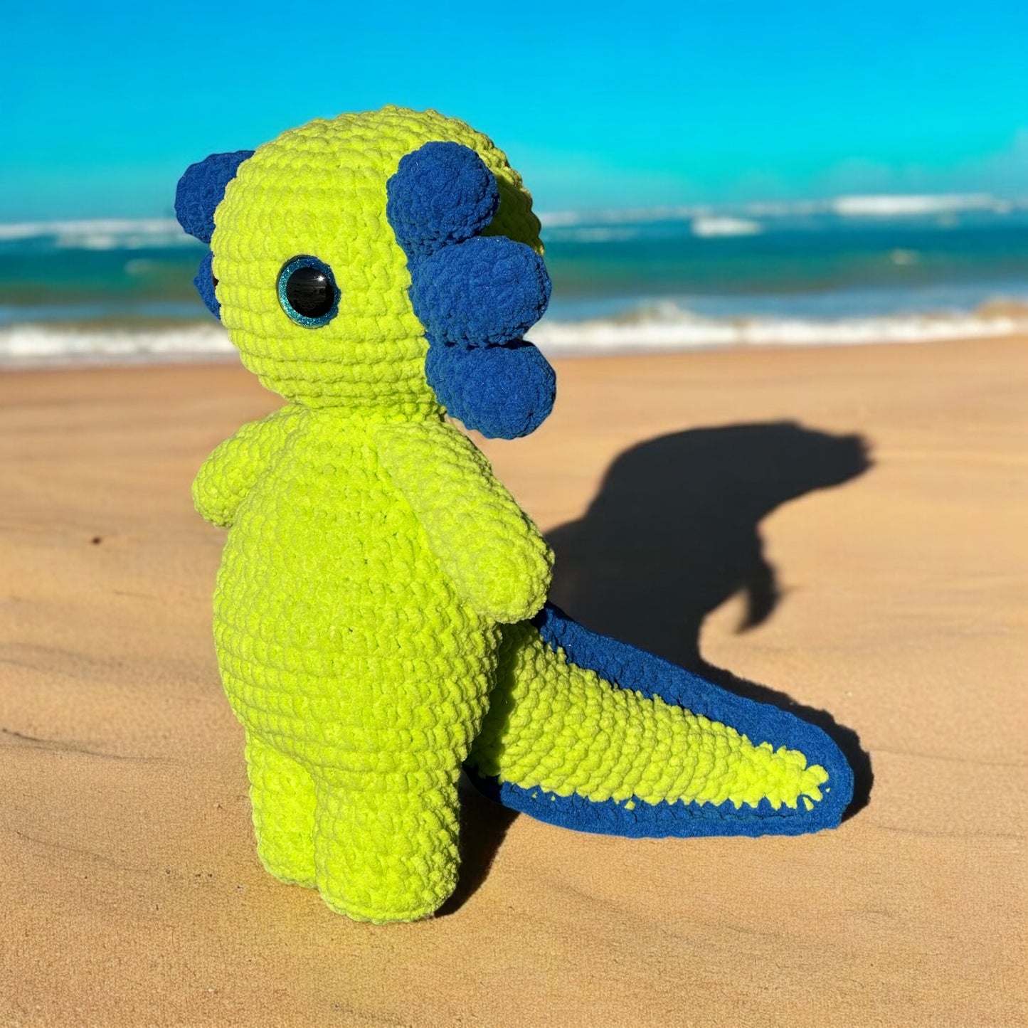 Crochet axolotl plush in bright neon yellow yarn with royal blue gill fronds and black safety eyes, photographed standing on beach sand with ocean waves in background