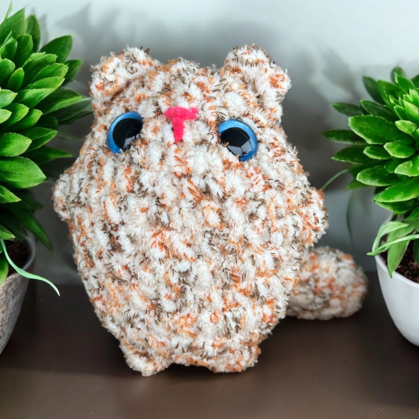 Chunky crochet kitten plush in speckled white and tan yarn with bright blue safety eyes and pink nose, photographed between green potted plants on dark surface