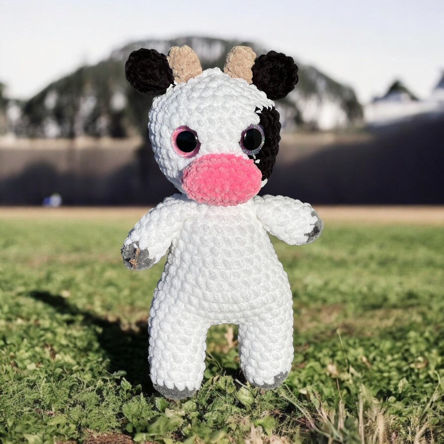 Crochet cow plush in white yarn with black and beige spot pattern on ears, pink nose, and safety eyes, photographed standing on grass with mountains in background