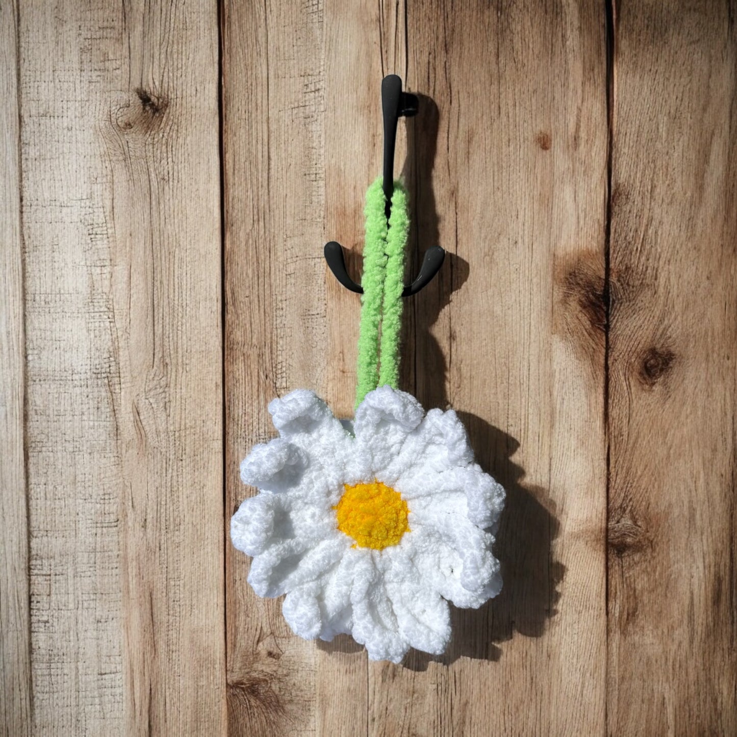Handmade crochet daisy with white petals and a yellow center, hanging from a black hook on a rustic wooden background. The flower has a soft, textured appearance with a green loop for hanging.