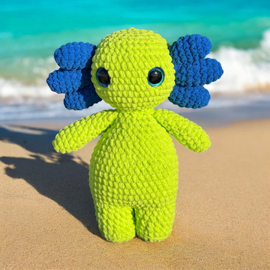 Side view of crochet axolotl plush in neon yellow with royal blue gills and tail fin, photographed on beach sand with shadow cast and ocean waves in background
