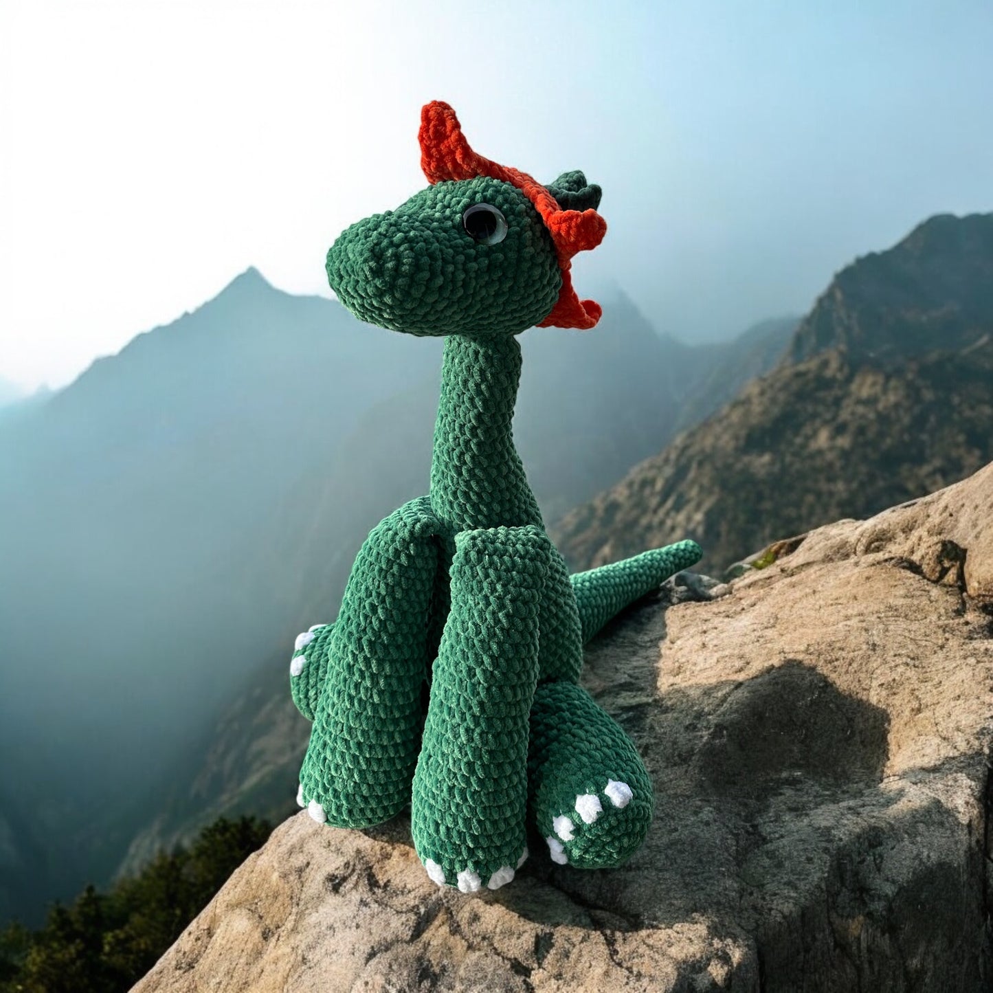Crochet Brontosaurus dinosaur in forest green yarn with orange spikes and white feet details, perched on rocky mountain ledge with dramatic misty mountain peaks in background
