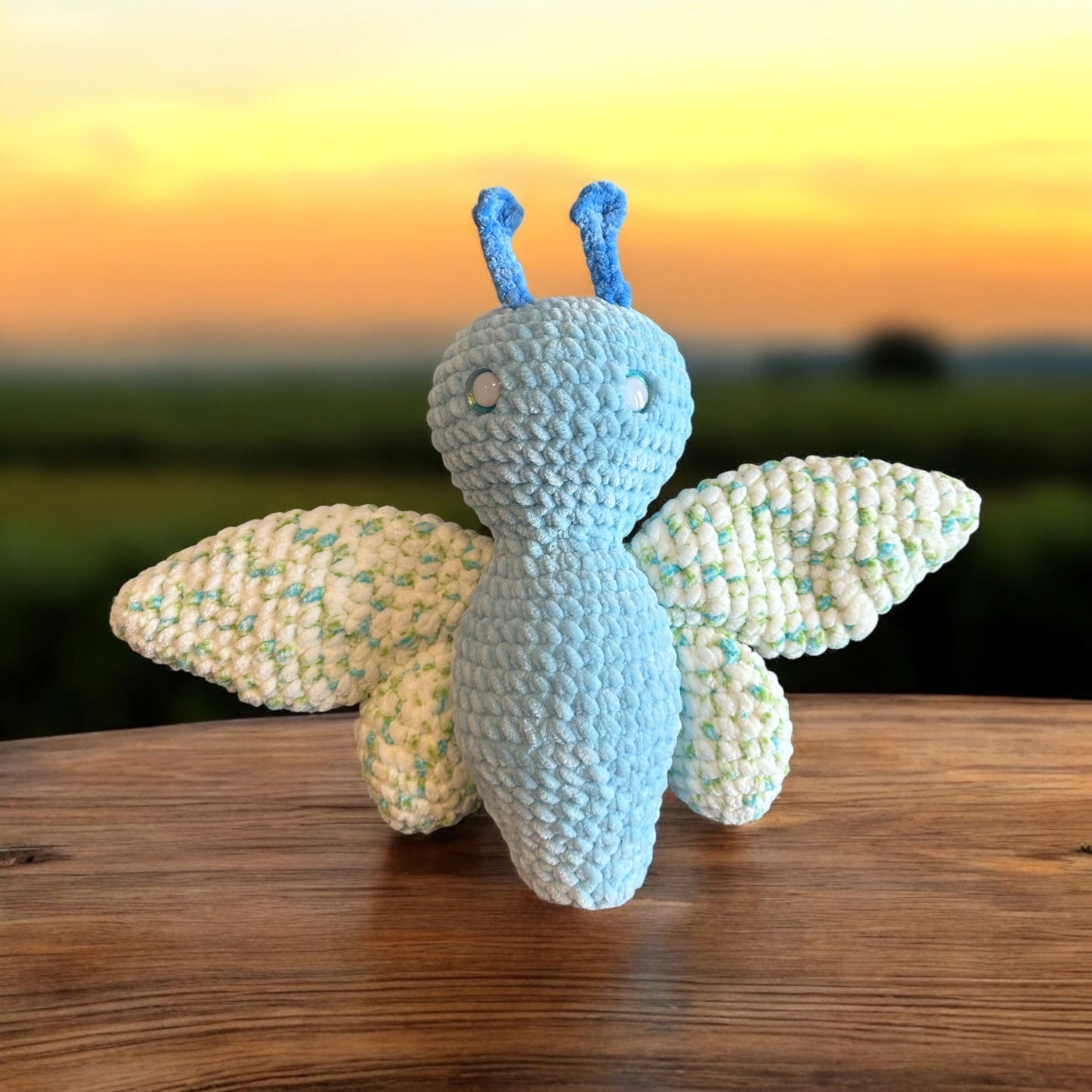 Crochet butterfly plush in soft white and light blue yarn with speckled pastel wing pattern, featuring blue antennae and safety eyes, photographed on wooden surface with sunset sky background