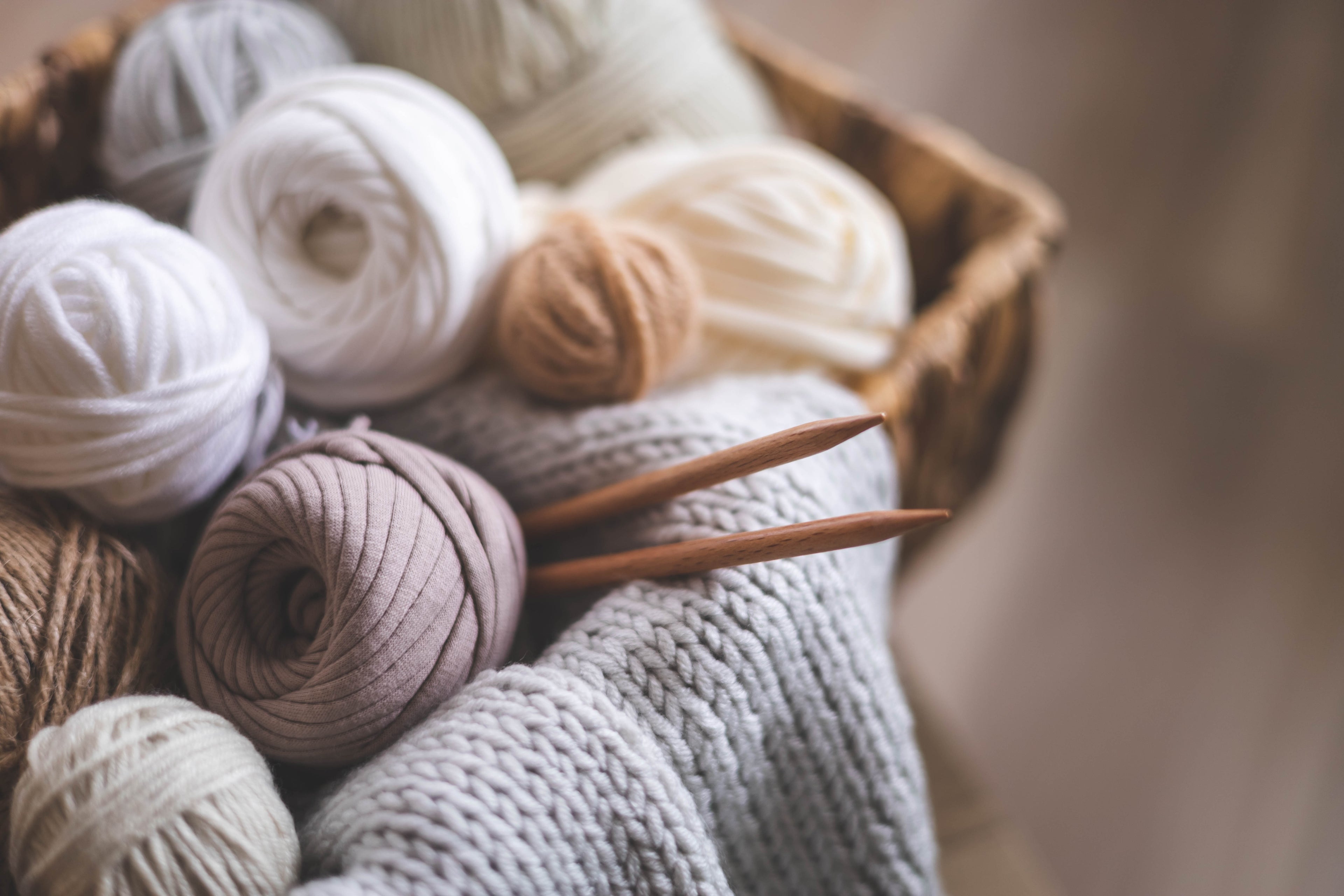 Cozy crochet supplies featuring wooden knitting needles resting on neutral-colored yarn balls in white, beige, and light gray, alongside a textured gray knitted piece in a wooden basket