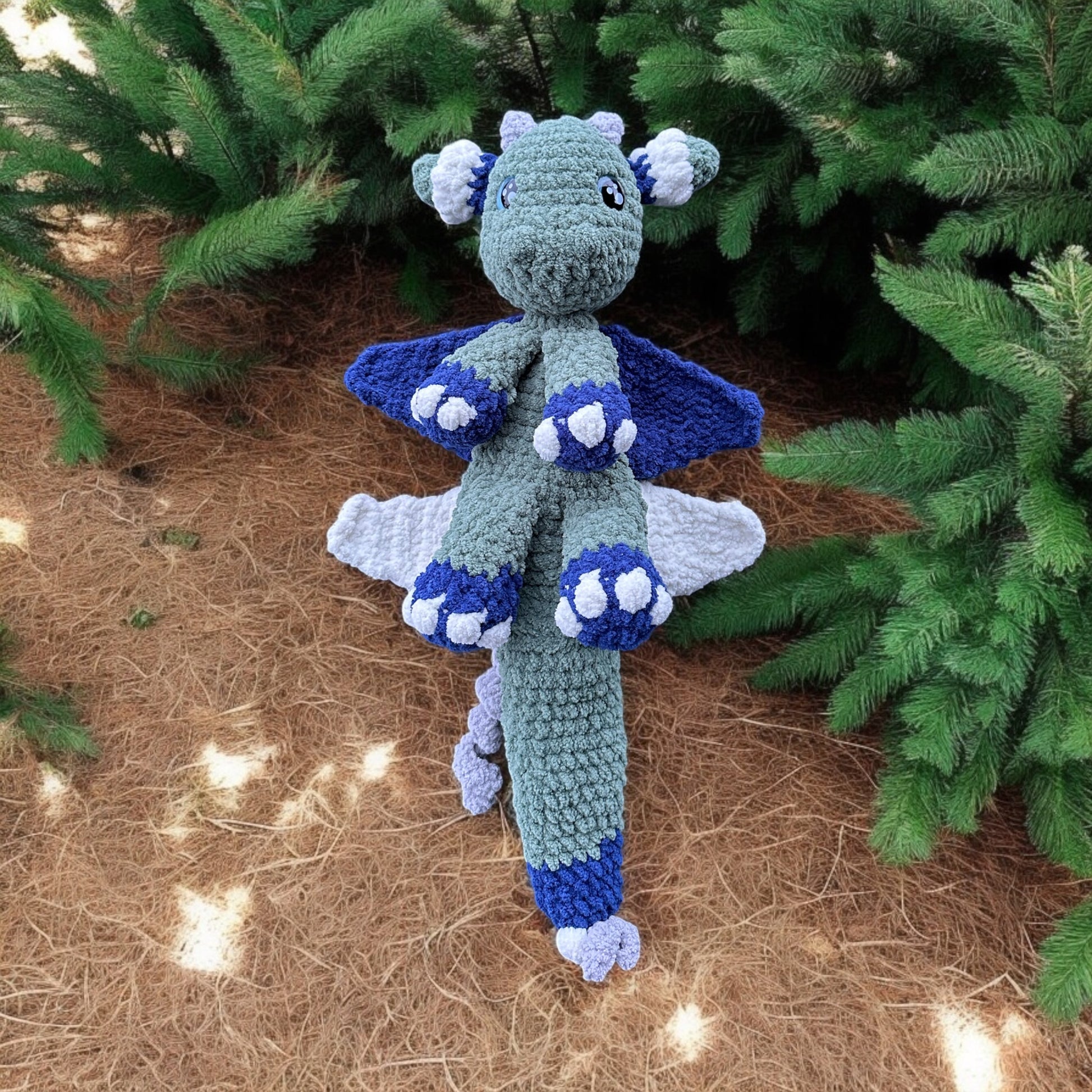 Crochet dragon snuggler in soft gray yarn with navy and white accents, spread out on brown pine needle ground with string lights and evergreen branches in background, featuring winged design and cozy paw details