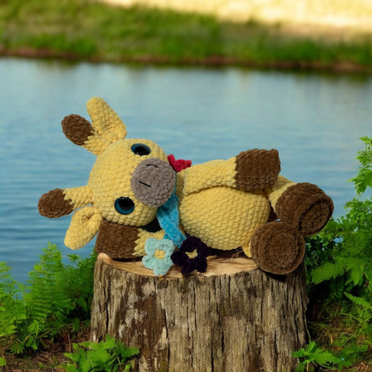 Crochet giraffe plush in yellow and brown yarn with blue safety eyes, wearing light blue bow tie and red hair bow, resting on tree stump with pond and ferns in background