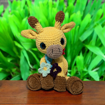Crochet giraffe plush in yellow and brown yarn sitting upright on wooden surface, with blue safety eyes, light blue bow tie and floral accents, photographed against vibrant green foliage background
