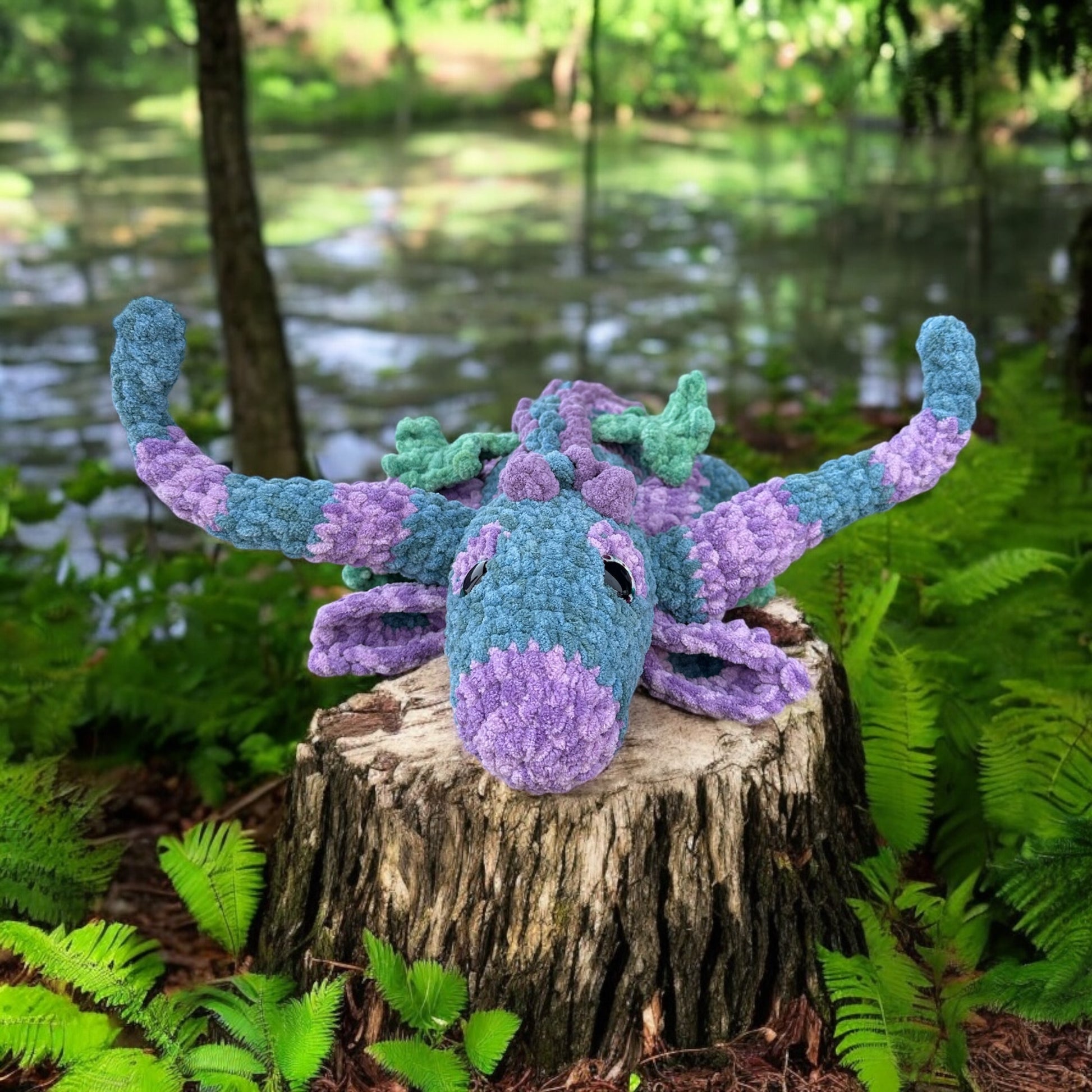Crochet dragon named Hooky in soft blue and purple textured yarn with green accents, photographed on tree stump surrounded by ferns and pond in woodland setting