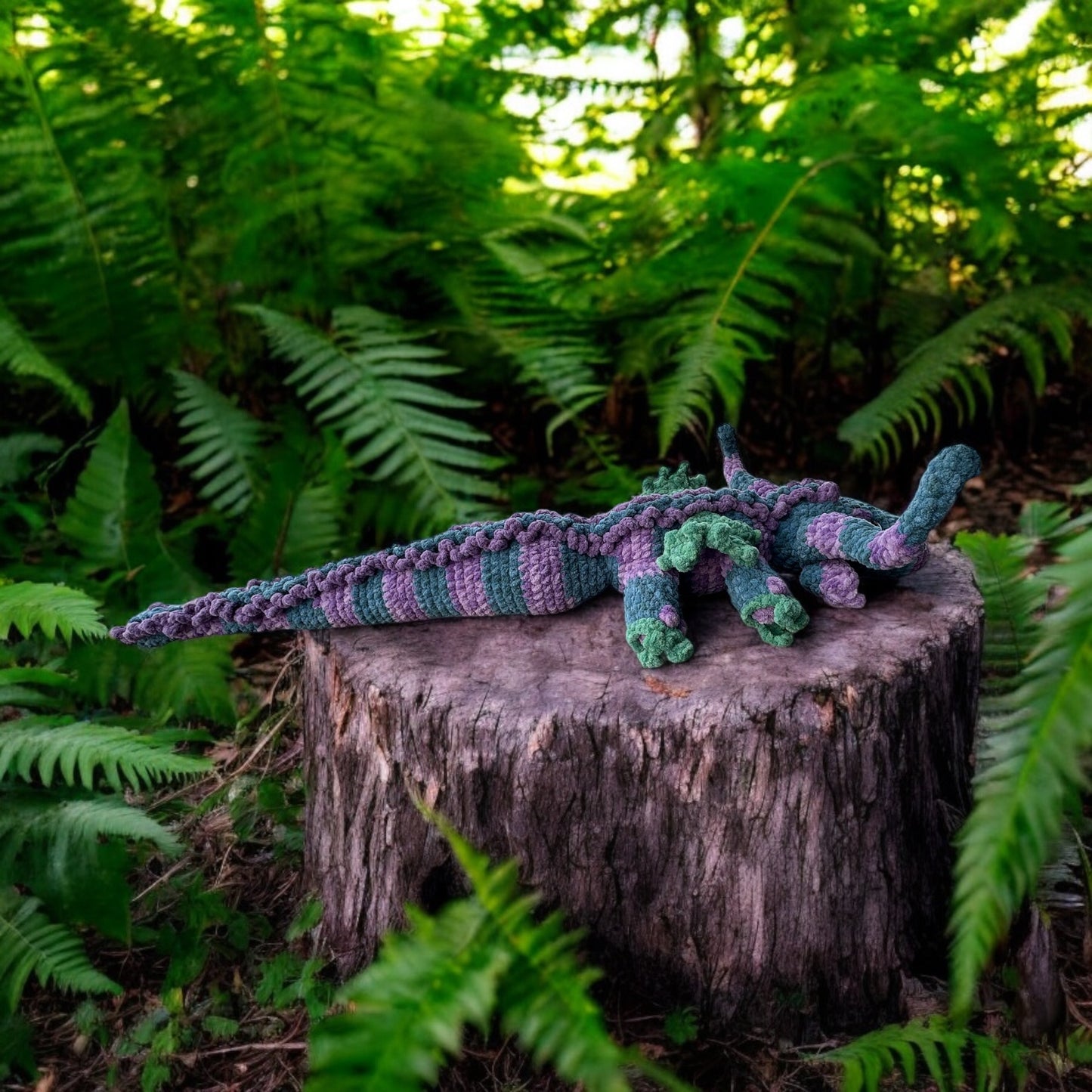 Crochet dragon named Hooky in soft blue and purple textured yarn with green accents, photographed on tree stump surrounded by ferns and sitting on a log.