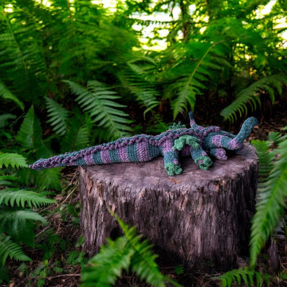 Crochet dragon named Hooky in soft blue and purple textured yarn with green accents, photographed on tree stump surrounded by ferns and sitting on a log.
