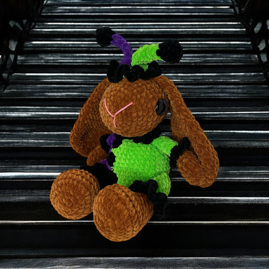 Plush brown crochet bunny wearing green and purple accessories photographed against black background