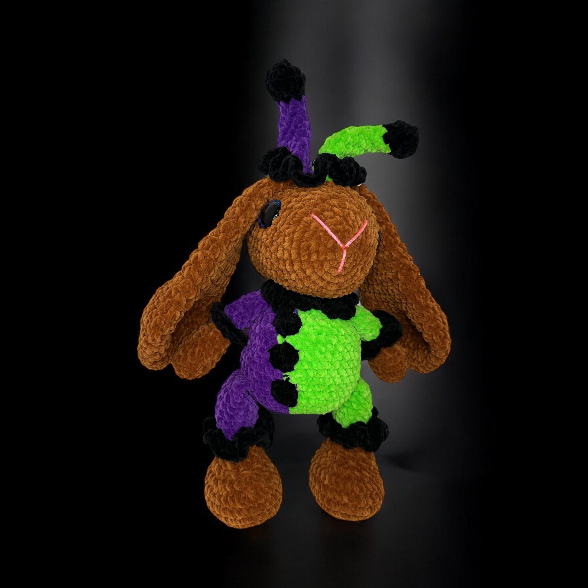 Plush brown crochet bunny wearing green and purple accessories photographed against a dark background