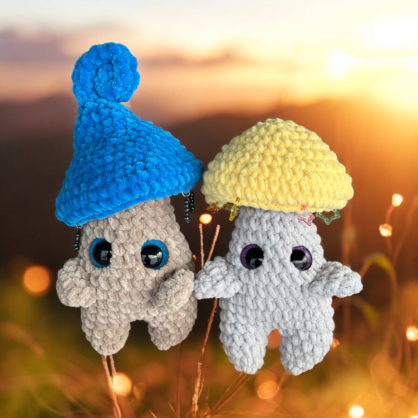 Two crochet mushroom sprites in soft gray and white yarn with blue and yellow mushroom caps, featuring safety eyes and tiny floral details, photographed against a warm sunset bokeh background