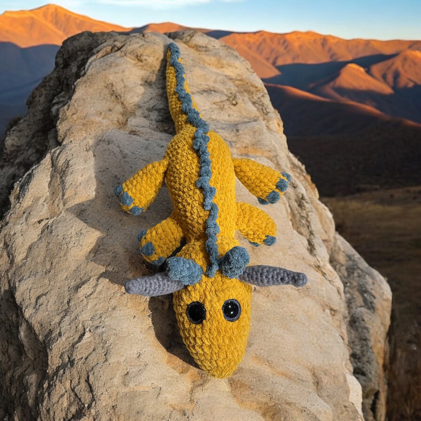 Crochet dragon named Patty in bright yellow and blue yarn with black safety eyes, photographed climbing forward on light colored rock formation with sunset-lit mountains in background