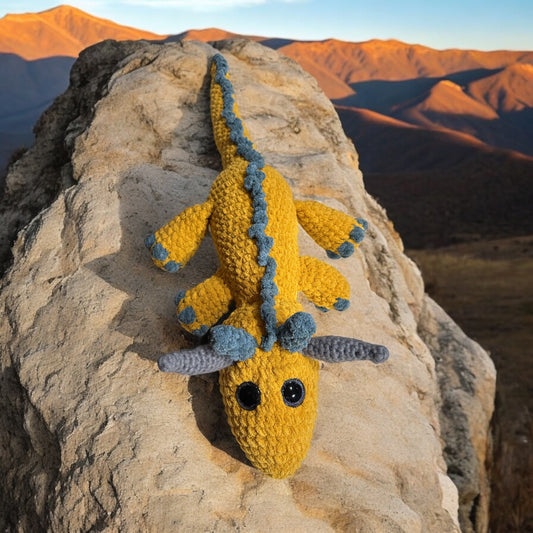 Crochet dragon named Patty in bright yellow and blue yarn with black safety eyes, photographed climbing forward on light colored rock formation with sunset-lit mountains in background