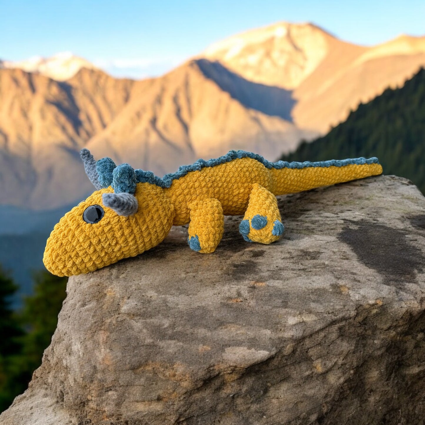 Crochet dragon named Patty in yellow yarn with blue accents and spikes, lying sideways on rocky ledge with golden sunlit mountain peaks in background