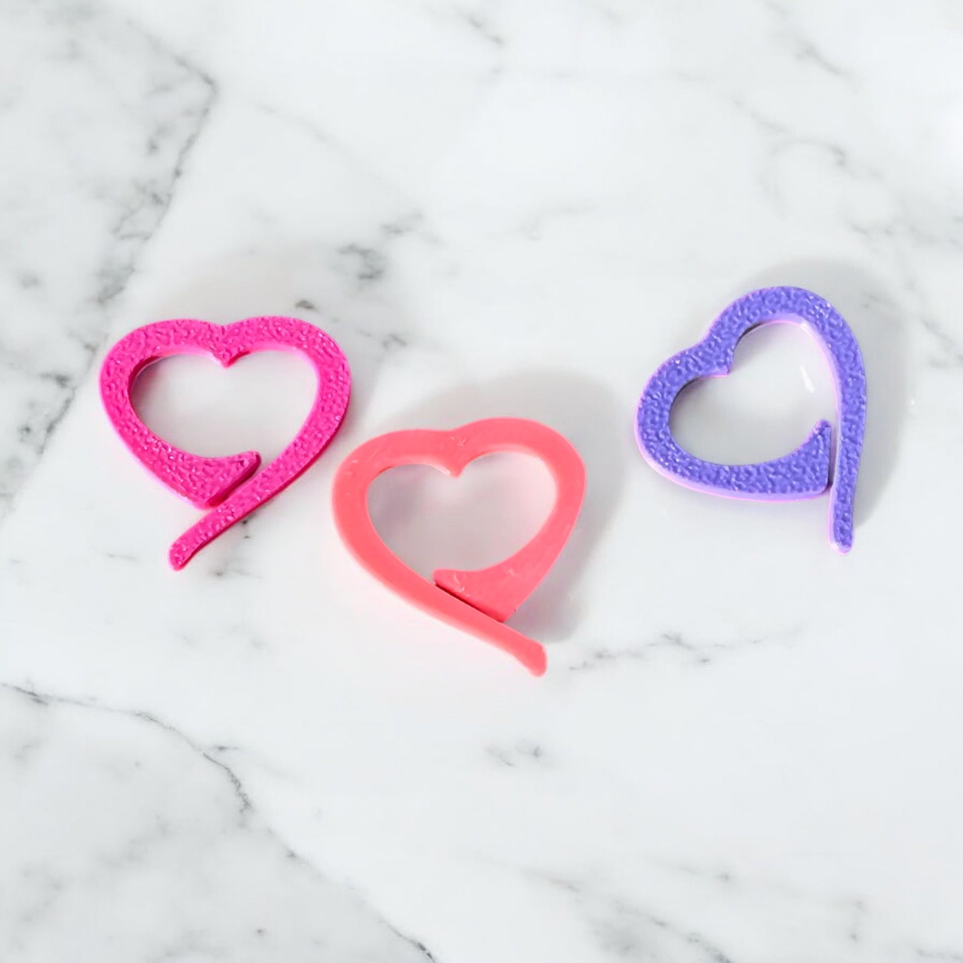 Three heart-shaped crochet stitch markers in pink, coral, and purple, arranged on white marble background, featuring textured finish and open heart design
