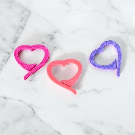 Three heart-shaped crochet stitch markers in pink, coral, and purple, arranged on white marble background, featuring textured finish and open heart design