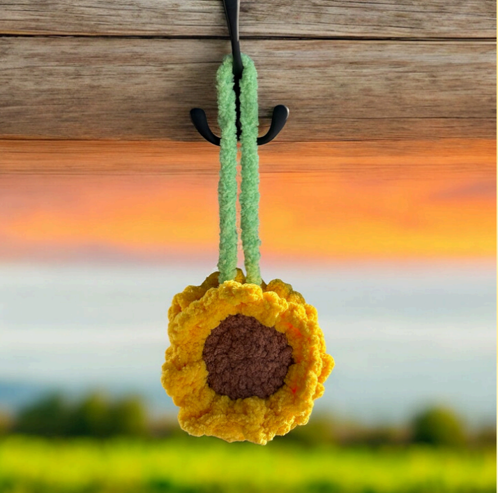Crochet sunflower car hanging ornament with bright yellow petals, brown center, and light green stem, photographed hanging against a sunset sky and green field background.