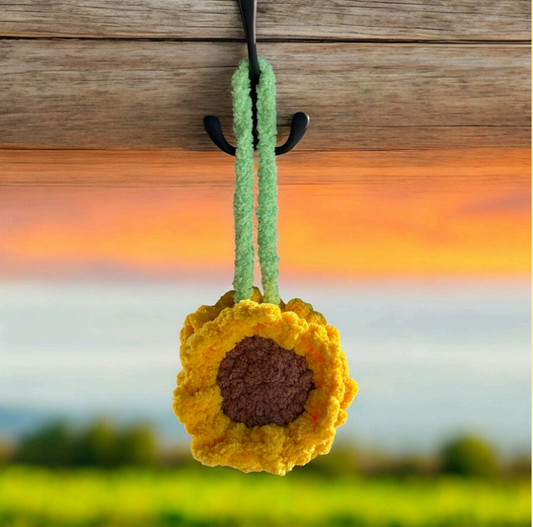 Crochet sunflower car hanging ornament with bright yellow petals, brown center, and light green stem, photographed hanging against a sunset sky and green field background.
