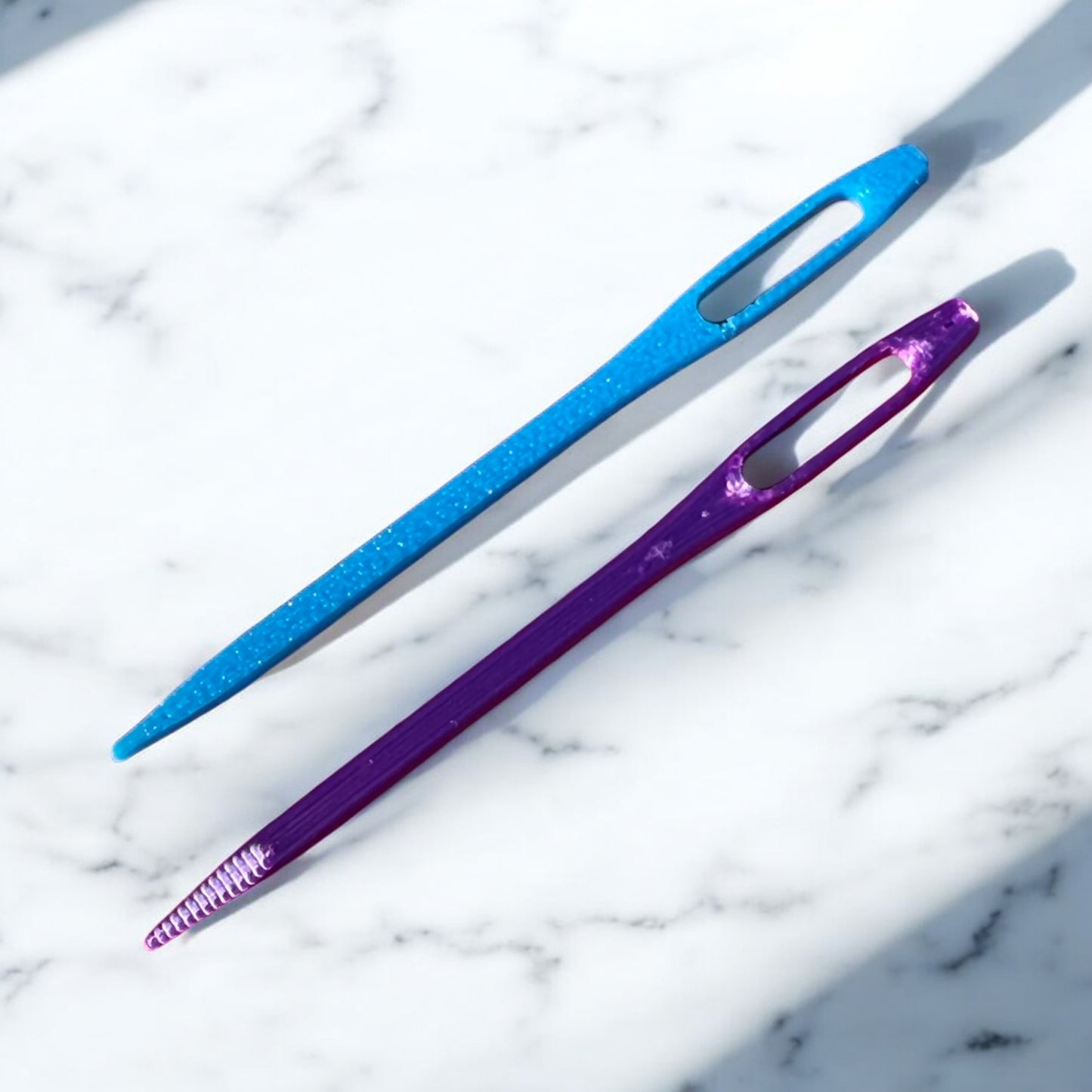 Two glitter finish yarn needles on white marble background - one metallic blue and one metallic purple with textured grip handles